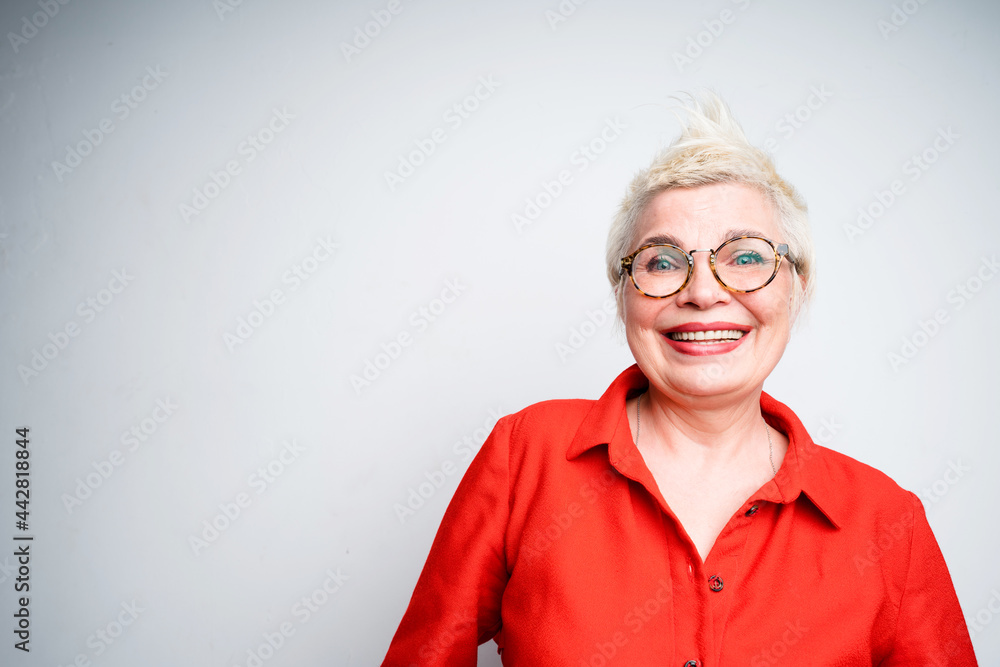 Cheerful elderly woman with mohawk wearing glasses and in a red shirt smiling with hands behind her back