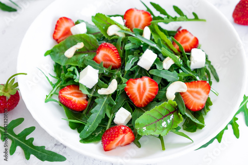 Tasty salad of strawberry with arugula, young beet tops, feta cheese and cashew nuts in white plate on marble table with salad leaves and fresh strawberry fruits around. Fruit detox and antioxidants.
