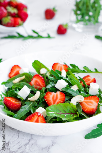 Vitamin salad of strawberry with arugula, spinach leaves, feta cheese and cashew nuts on marble table with salad leaves and fresh strawberry fruits around. Vegan food, keto diet and paleo diet.