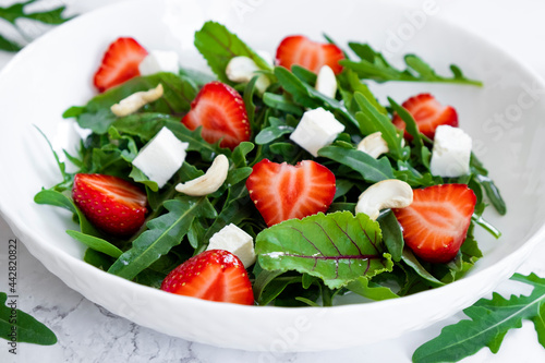 Vitamin salad of strawberry with arugula, spinach leaves, feta cheese and cashew nuts in white plate with salad leaves and fresh strawberry fruits around. Vegan food, keto diet and paleo diet.