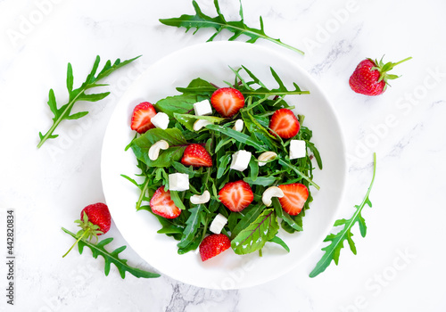 Summer salad of strawberry with arugula, spinach leaves, feta cheese and cashew nuts in white plate on marble table with salad leaves and fresh strawberry fruits around. Raw food diet.
