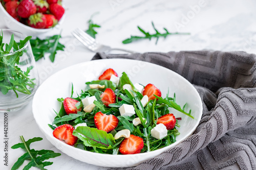 Summer salad of strawberry with arugula, spinach leaves, feta cheese and cashew nuts in white plate with gray towel, salad leaves and fresh strawberry fruits around. Vegetarian food and raw food diet.