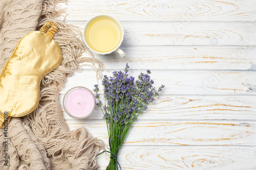 Cozy composition with warm blanket, herbal tea, aromatic candle, bouquet of lavender and sleeping mask on white wooden background. Insomnia and sleep optimization concept. Hygge style composition.