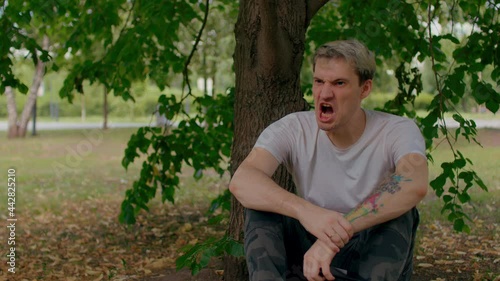 Young man rests, sitting near tree trunk in parkland. Guy burps and smiles, looking away and sitting near tree in summertime
