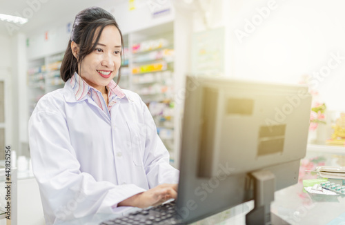 smart asian female pharmacist typing drug list with computer in drugstore, she feeling happy and smile, she holding hand on keyboard on counter
