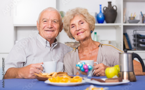 Smiling elderly spouses enjoying time at home, drinking tea with sweets