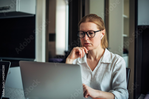 Adult 40-year-old woman in glasses and white shirt is working with laptop in a home office. Positive female writer. Freelance, coworking, remote work, self-employment, lockdown.