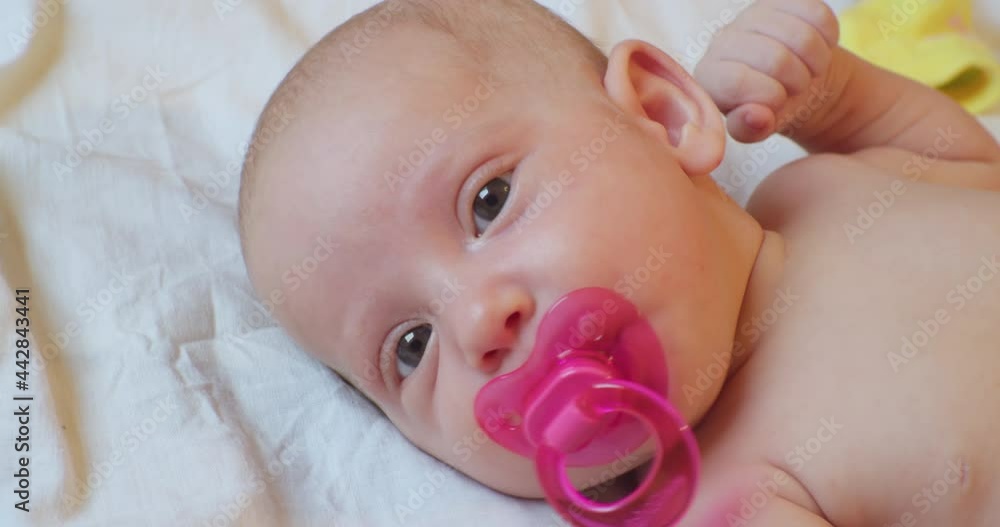 cute baby infant with pink pacifier in mouth lies in bed. portrait. close-up. cute baby infant with pink pacifier in mouth lies in bed. portrait. close-up.