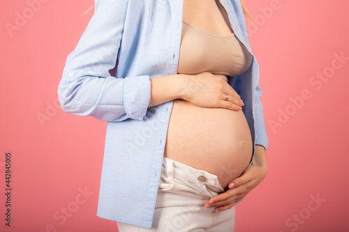 Beautiful pregnant woman in a blue shirt stroking her belly on a pink background.