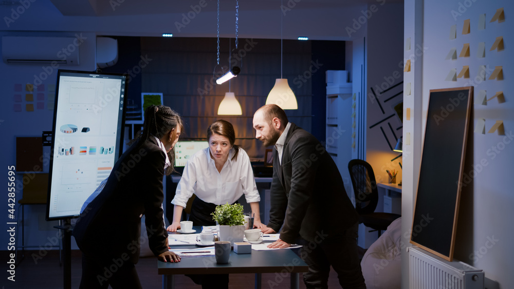 Diverse multi ethnic teamwork standing around table brainstorming company ideas checking management statistics paperwork. Focused coworkers planning overtime business presentation in meeting room Diverse multi ethnic teamwork standing around table brainstorming company ideas checking management statistics paperwork. Focused coworkers planning overtime business presentation in meeting room
