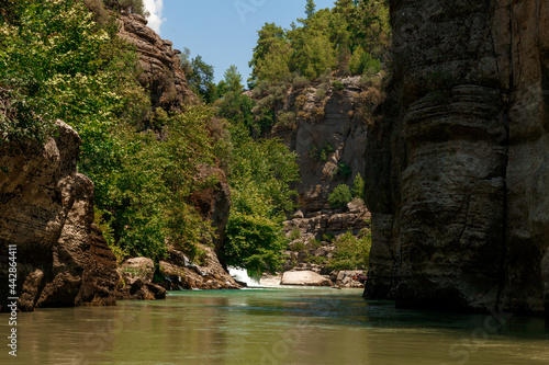 A rocky cliff with green trees and bushes and the surface of a mountain river in a canyon with a small foamy waterfall on a background