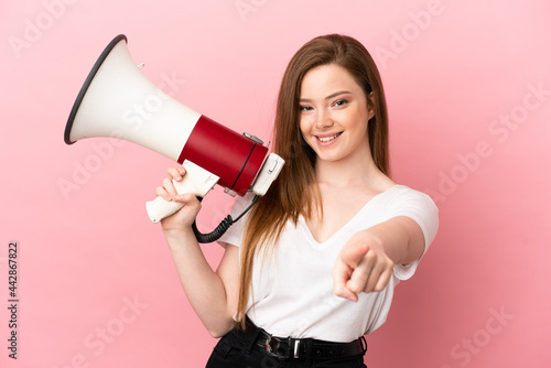 Teenager girl over isolated pink background holding a megaphone and smiling while pointing to the front