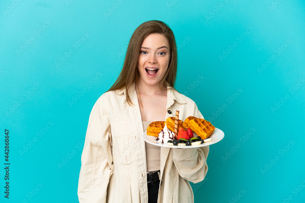 Teenager girl holding waffles over isolated blue background with surprise facial expression