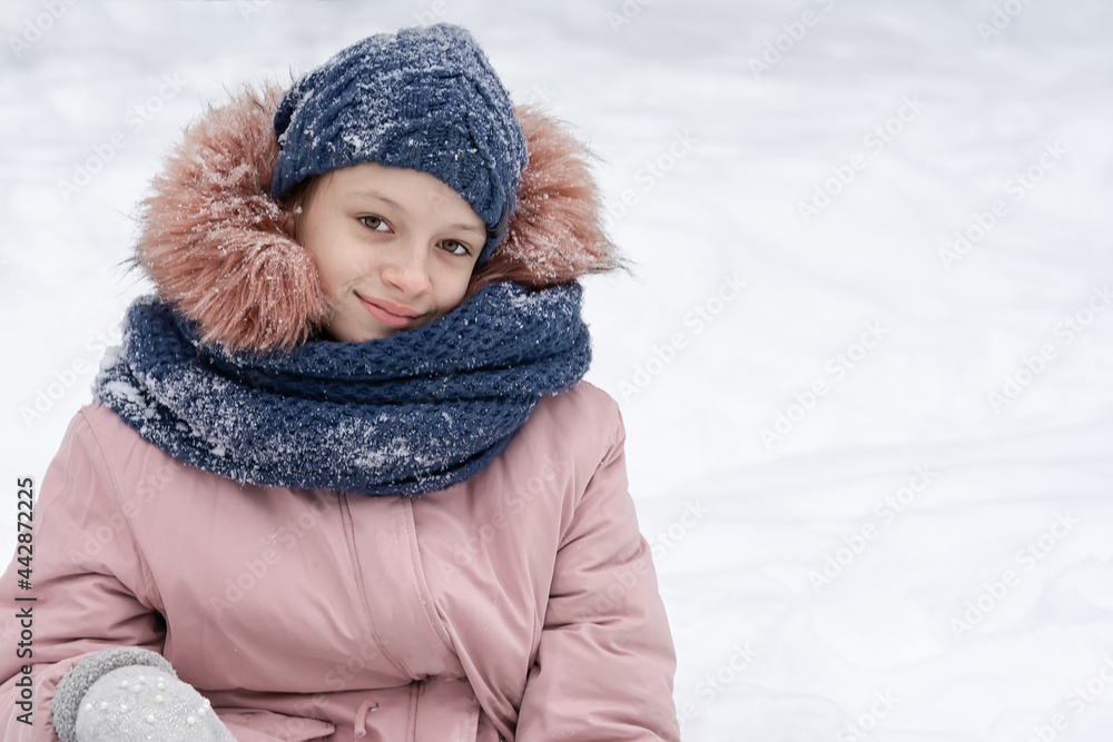 A teenage girl in a snowy blue hat and a pink jacket sits in the snow and smiles. People, lifestyle concept A teenage girl in a snowy blue hat and a pink jacket sits in the snow and smiles. People, lifestyle concept