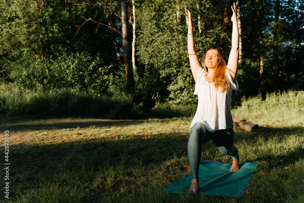Sporty young woman practicing yoga, outdoors. Yogi girl standing on a sports mat in yoga asana near the forest, outside. Exercise and meditation in nature. Healthy active lifestyle concept