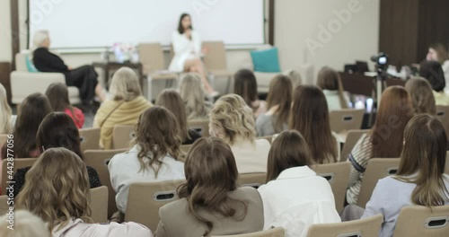 The female audience is listening to the speaker in the small cozy conference room. There are two women on the stage. There is a camera in the aisle. Back view. Education concept.