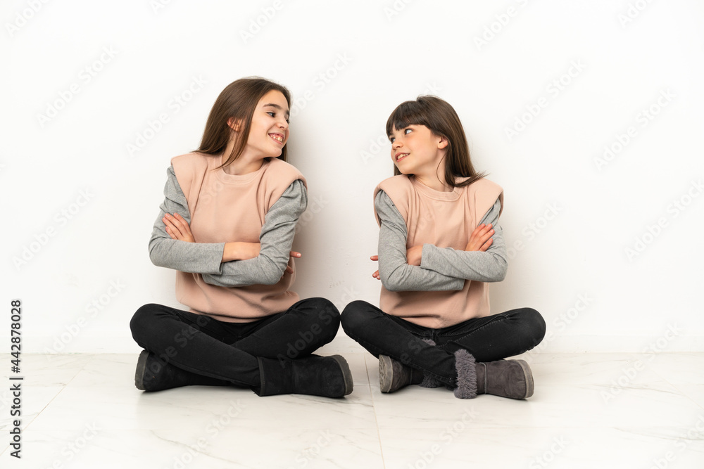Little sisters sitting on the floor isolated on white background looking over the shoulder with a smile Little sisters sitting on the floor isolated on white background looking over the shoulder with a smile