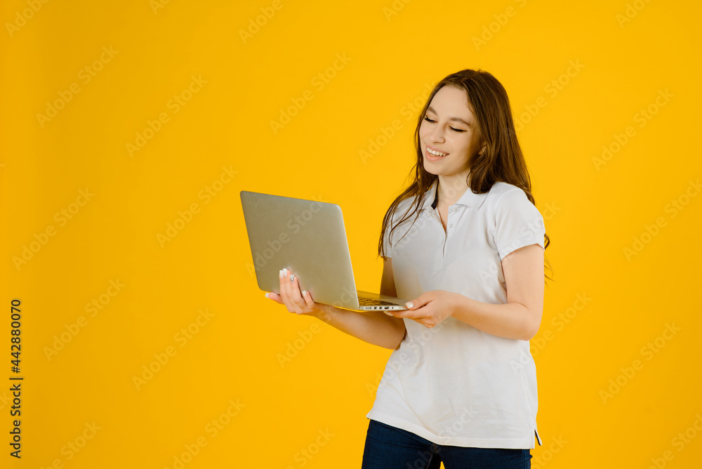 Portrait of a young happy woman in white t-shirt smiling and holding a notebook computer on a yellow background.