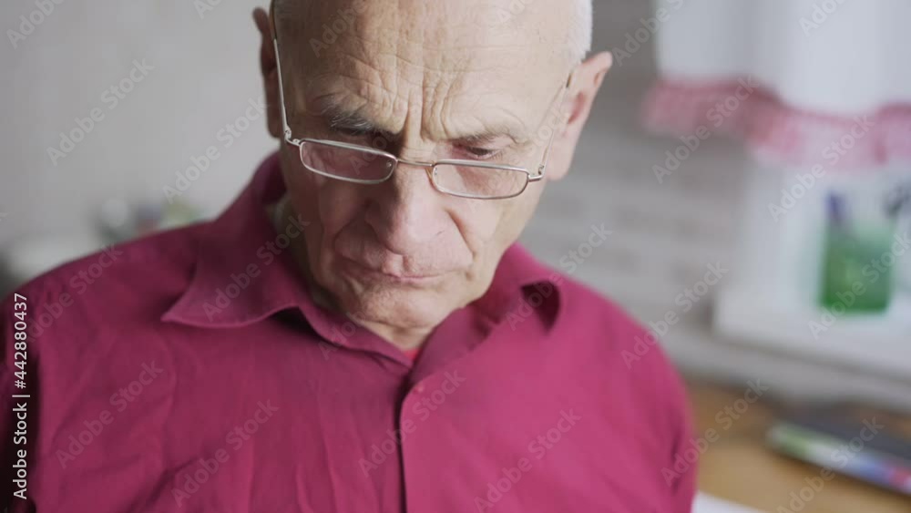 Pensioner man with grey hair and glasses reads large sheet of newspaper sitting in kitchen under electric light close view Pensioner man with grey hair and glasses reads large sheet of newspaper sitting in kitchen under electric light close view