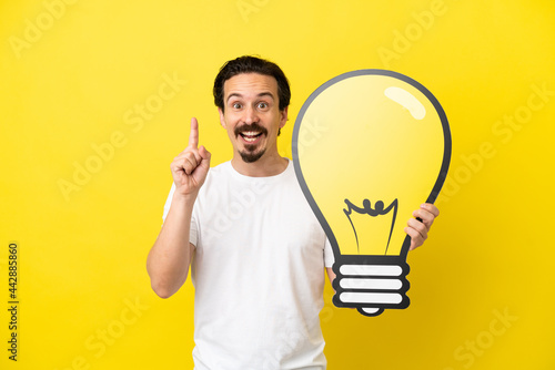 Young caucasian man isolated on yellow background holding a bulb icon and thinking