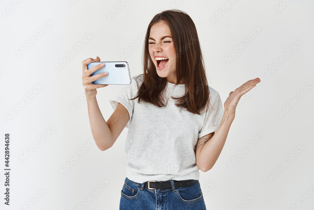 Happy woman laughing and looking at smartphone screen, watching funny video, standing over white background Happy woman laughing and looking at smartphone screen, watching funny video, standing over white background