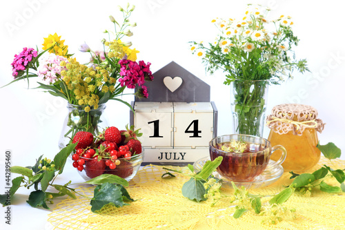 Calendar for July 14 : the name of the month of July in English, cubes with the number 14, bouquets of wild flowers, jam, fruit, a cup of tea on a yellow openwork napkin © MARYIA