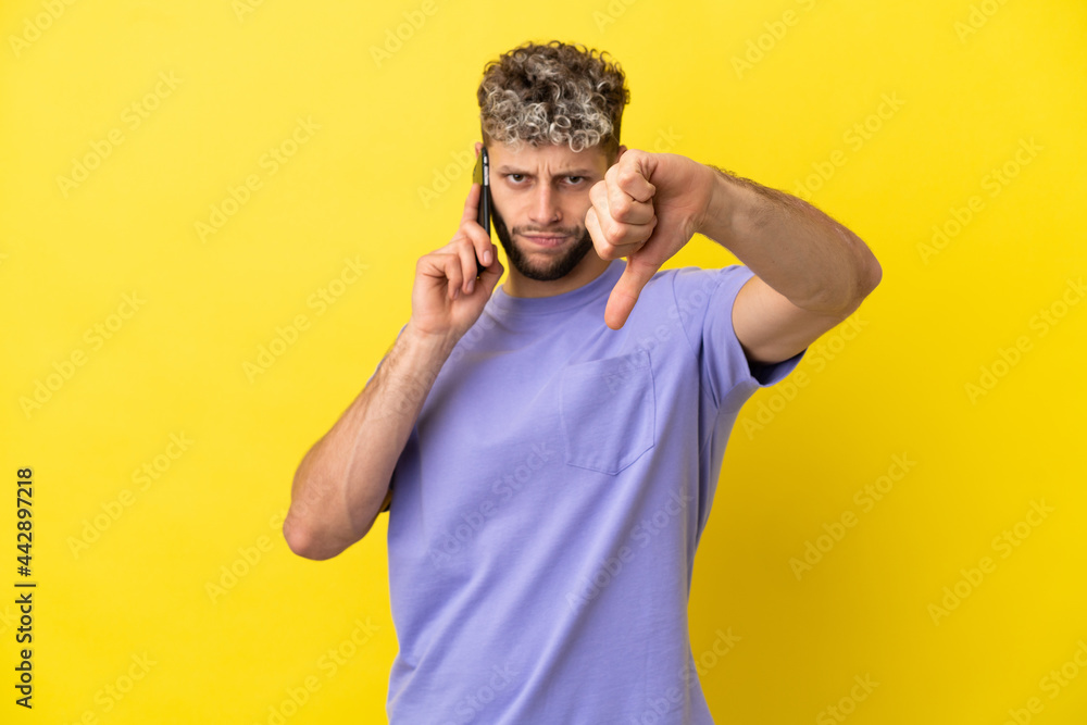 Young caucasian man using mobile phone isolated on yellow background showing thumb down with negative expression Young caucasian man using mobile phone isolated on yellow background showing thumb down with negative expression