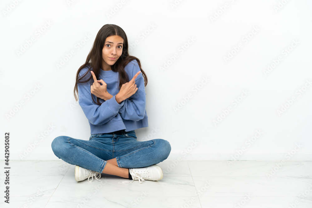Young French girl sitting on the floor pointing to the laterals having doubts Young French girl sitting on the floor pointing to the laterals having doubts