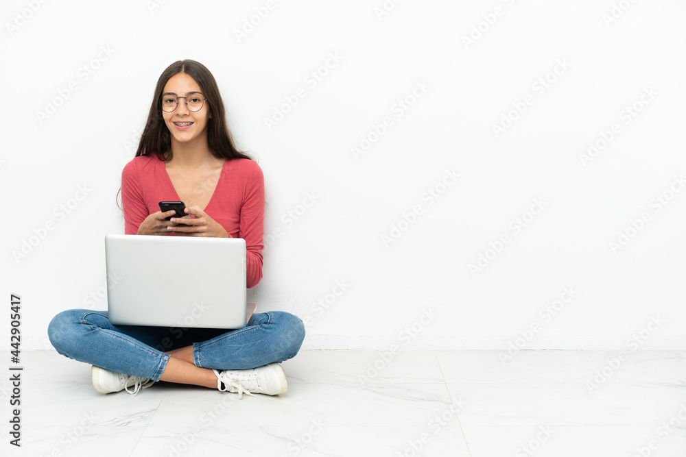 Young French girl sitting on the floor with her laptop sending a message with the mobile Young French girl sitting on the floor with her laptop sending a message with the mobile