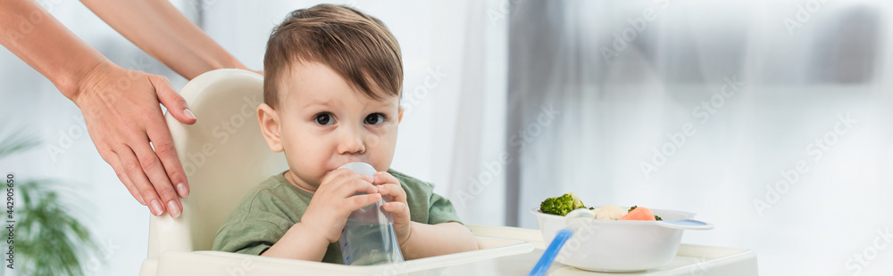 Mother holding high chair near toddler son with baby bottle and food, banner Mother holding high chair near toddler son with baby bottle and food, banner