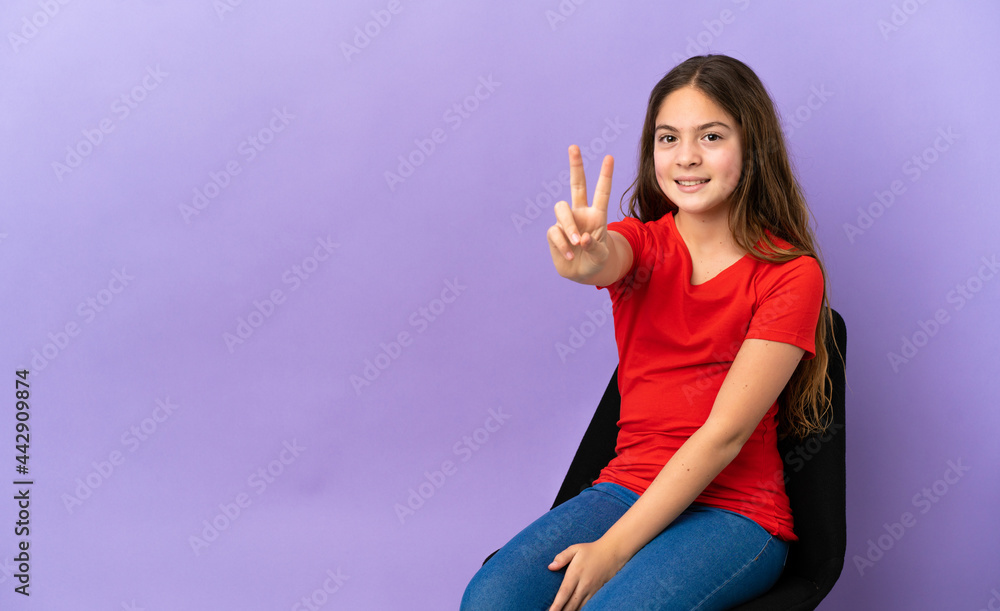 Little caucasian girl sitting on a chair isolated on purple background smiling and showing victory sign Little caucasian girl sitting on a chair isolated on purple background smiling and showing victory sign