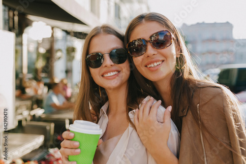 Close up portrait of two adorable girls wearing sunglasses looking at camera and smiling while walking and drinking coffee in sunlight 