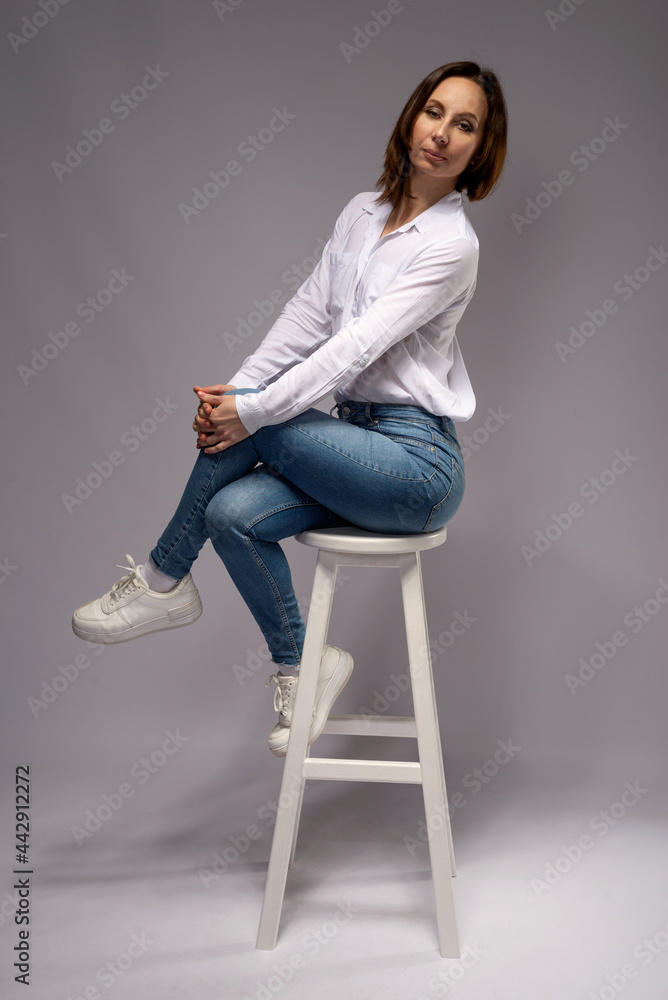Stylish woman in jeans and white shirt posing on a bar stool, gray background. Stylish woman in jeans and white shirt posing on a bar stool, gray background.