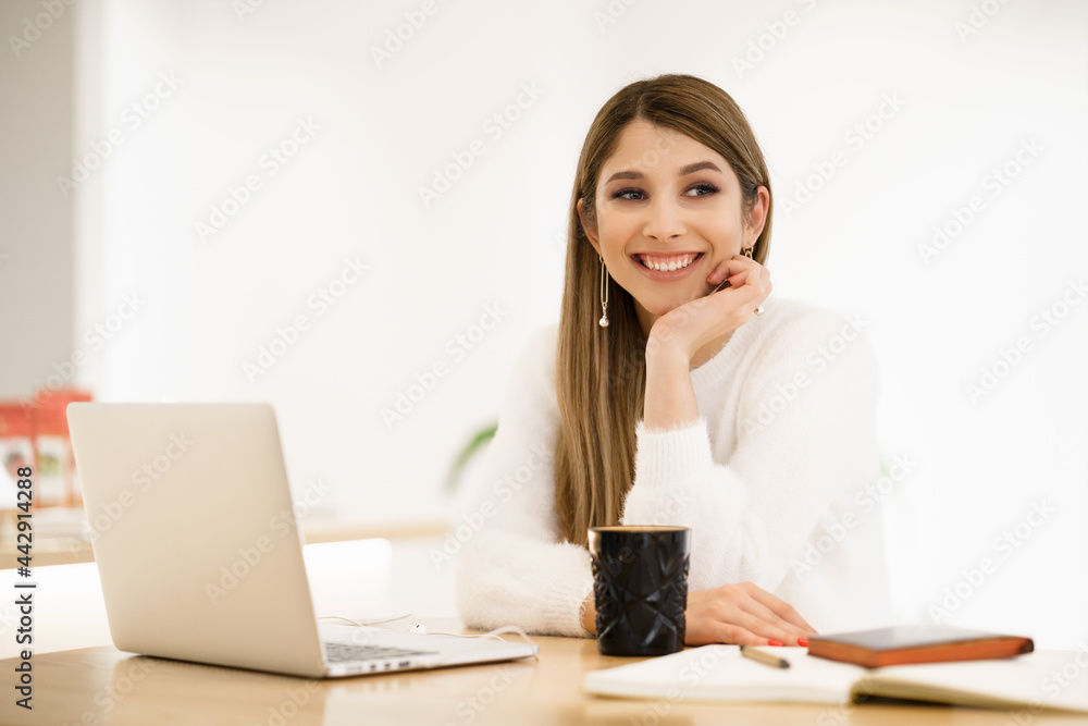 Beautiful young woman in casual clothing using laptop and smiling while working indoors Beautiful young woman in casual clothing using laptop and smiling while working indoors