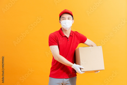 Young delivery man in medical mask holding and carrying a cardboard isolated on yellow background