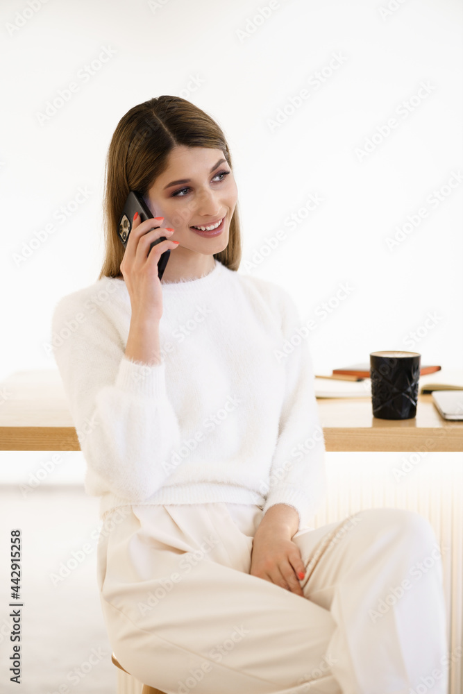 Woman with candid smile chatting by phone and enjoying coffee break in cafe . Wearing casual white sweater and pants. Woman with candid smile chatting by phone and enjoying coffee break in cafe . Wearing casual white sweater and pants.
