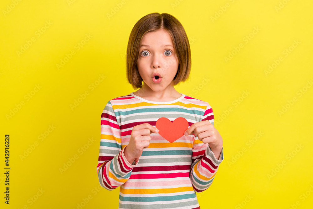 Photo of amazed shocked small girl hold hands small red paper heart shape isolated on yellow color background Photo of amazed shocked small girl hold hands small red paper heart shape isolated on yellow color background