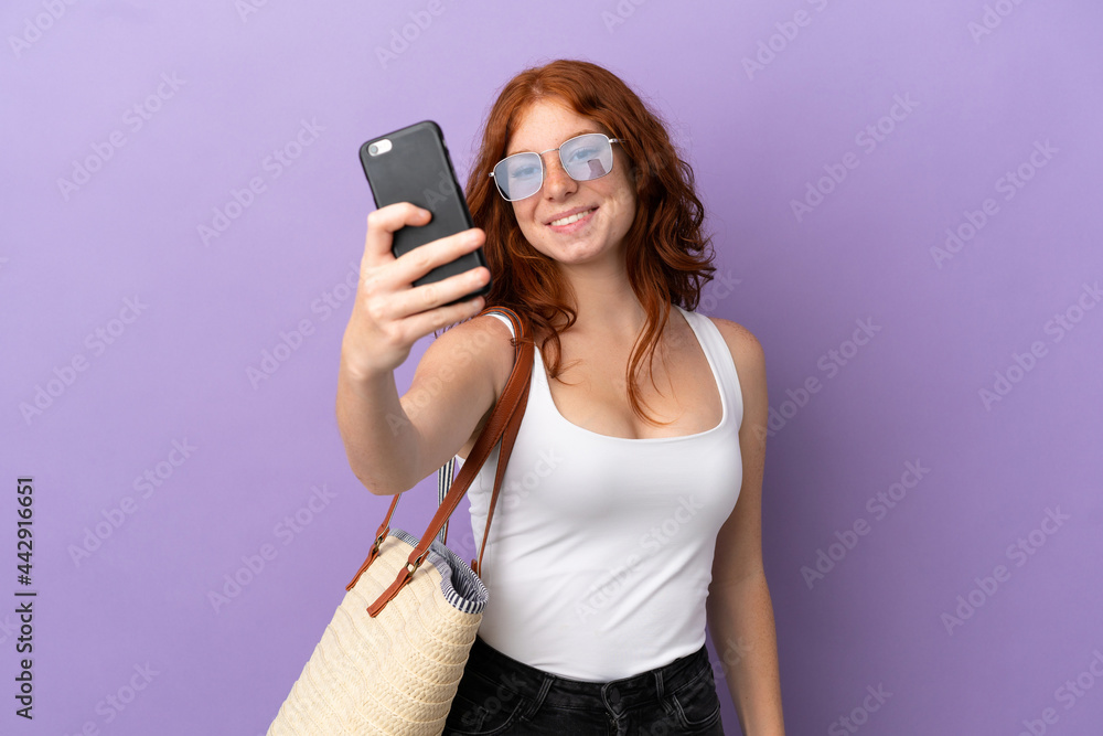 Teenager redhead girl holding a beach bag isolated on purple background making a selfie Teenager redhead girl holding a beach bag isolated on purple background making a selfie