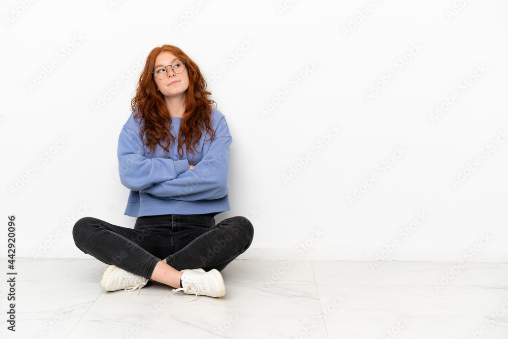 Teenager redhead girl sitting on the floor isolated on white background making doubts gesture while lifting the shoulders Teenager redhead girl sitting on the floor isolated on white background making doubts gesture while lifting the shoulders