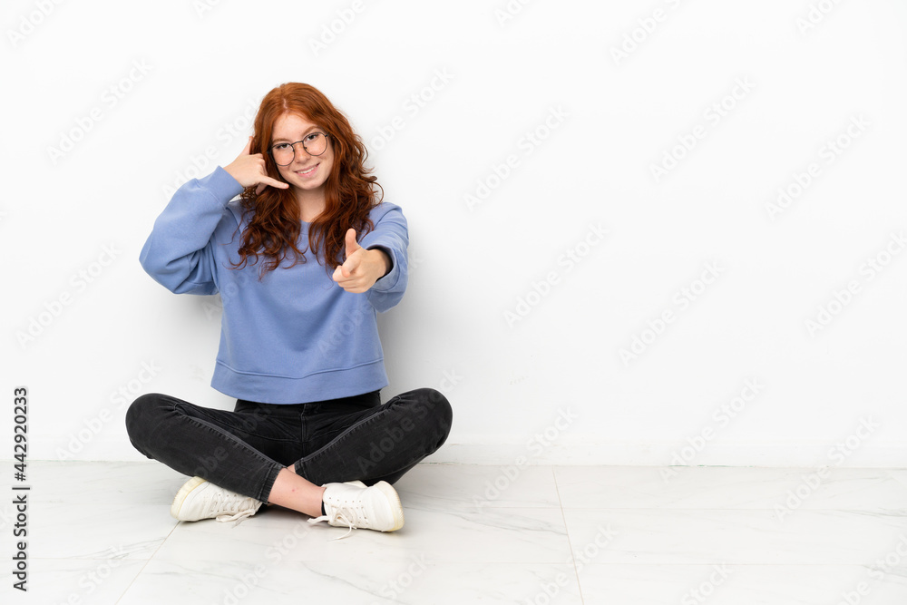Teenager redhead girl sitting on the floor isolated on white background making phone gesture and pointing front Teenager redhead girl sitting on the floor isolated on white background making phone gesture and pointing front