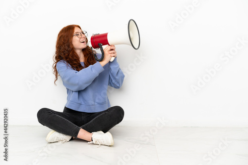 Teenager redhead girl sitting on the floor isolated on white background shouting through a megaphone
