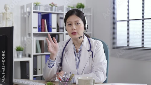 Wallpaper Mural medical worker looking at the camera and wearing headphones is explaining with her hand gesturing in a online video call at a bright clinic office. Torontodigital.ca