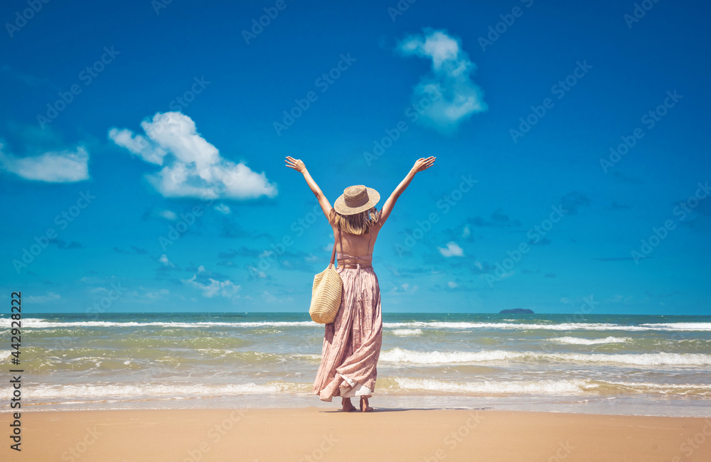 Happy traveller woman in sunhat and dress standing with her arms raised looking tropical beach on summer day Happy traveller woman in sunhat and dress standing with her arms raised looking tropical beach on summer day