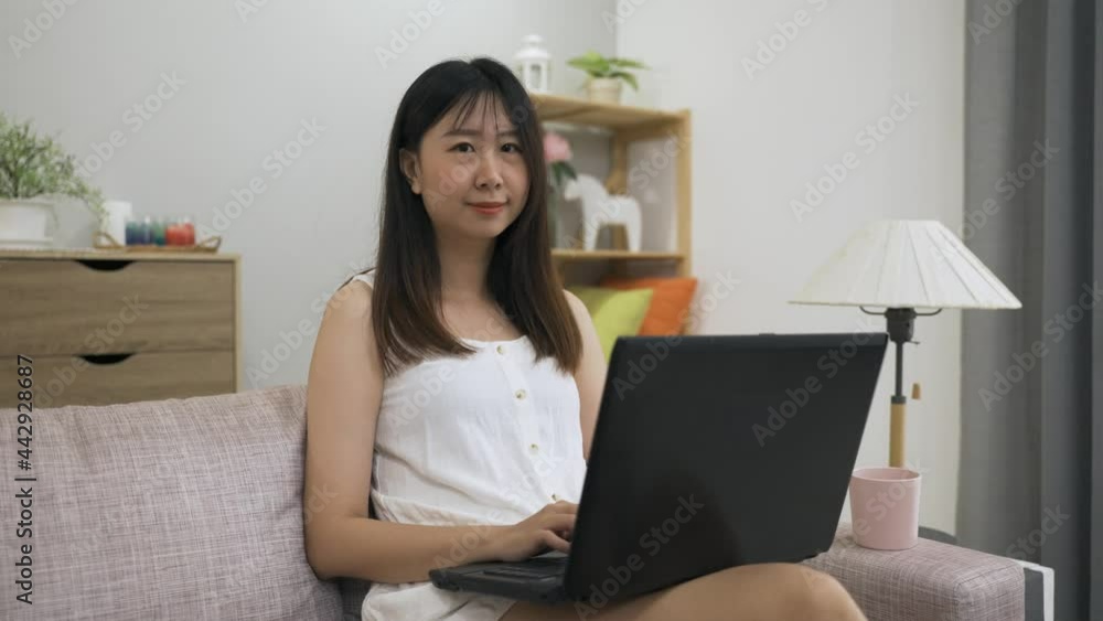 attractive asian female is turning to look at the camera with a smile while working from home on her laptop on the couch in a bright modern living room. attractive asian female is turning to look at the camera with a smile while working from home on her laptop on the couch in a bright modern living room.