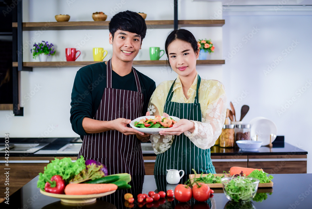 Asian couples in the apron are making shrimp salad for a holiday in the kitchen, with a stove pan, cutting board and ingredients such as Lettuce, green oak, red oak,tomatoes, carrot purple cabbage Asian couples in the apron are making shrimp salad for a holiday in the kitchen, with a stove pan, cutting board and ingredients such as Lettuce, green oak, red oak,tomatoes, carrot purple cabbage
