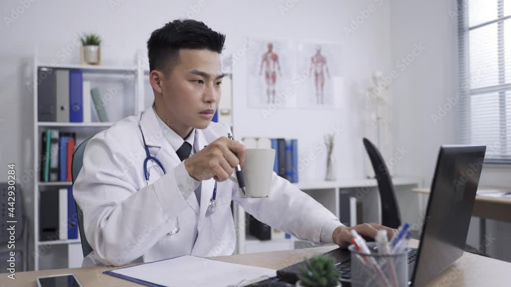 hardworking asian male medical worker is continuing with his work on the laptop after pausing to think while sipping tea from mug in a hospital office. hardworking asian male medical worker is continuing with his work on the laptop after pausing to think while sipping tea from mug in a hospital office.