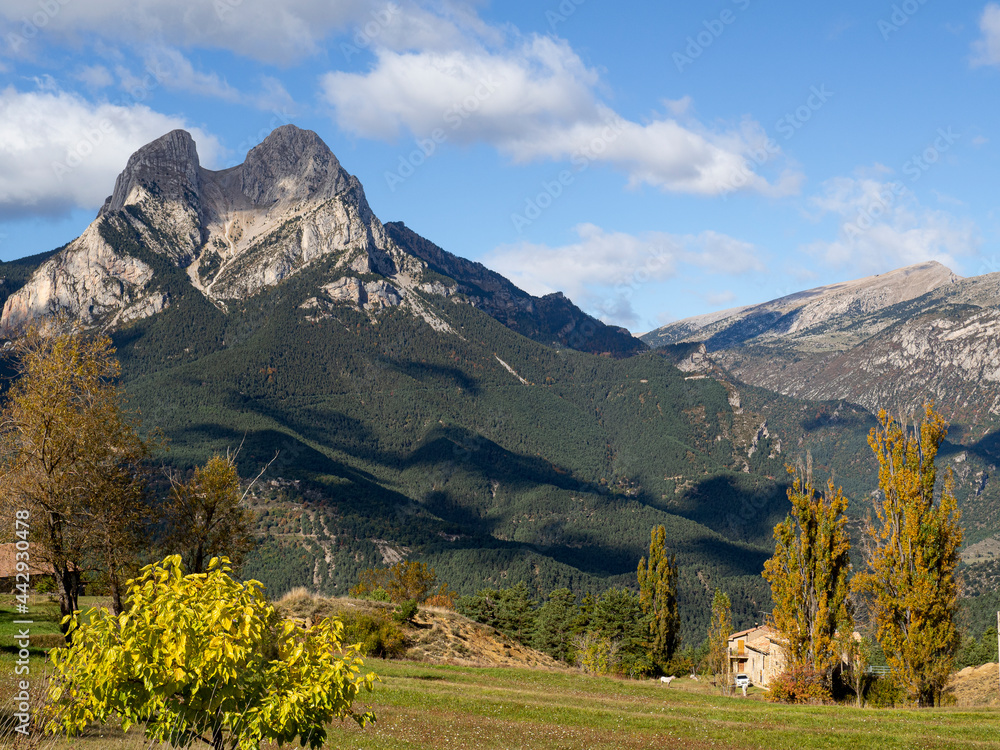 Montaña del Pedraforca en Cataluña, con una forma singular, sobre un cielo azul con nubes blancas sombreando el valle con árboles de colores de otoño, marrones y amarillos en Octubre de 2020.
