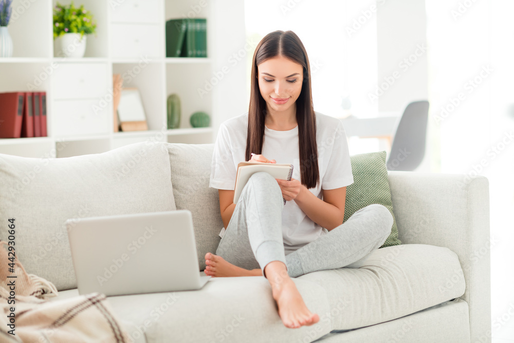 Photo of nice clever lady hold notepad write notes wear white t-shirt sit couch barefoot in room indoors Photo of nice clever lady hold notepad write notes wear white t-shirt sit couch barefoot in room indoors