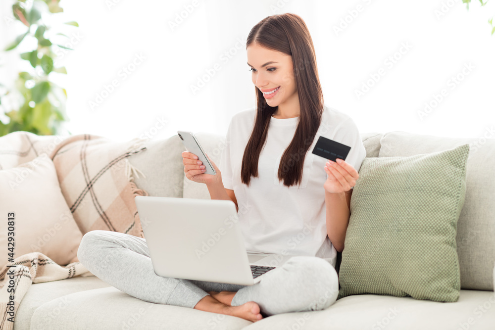 Photo of cheerful lady hold computer debit card texting phone nfc pay wear white t-shirt sit couch in room indoors Photo of cheerful lady hold computer debit card texting phone nfc pay wear white t-shirt sit couch in room indoors