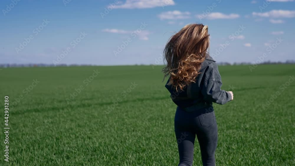 Girl is running on green field. Young woman with long fluttering hair in denim suit runs in nature and turning her cheerful face on camera. Rear view. Slow motion. Girl is running on green field. Young woman with long fluttering hair in denim suit runs in nature and turning her cheerful face on camera. Rear view. Slow motion.